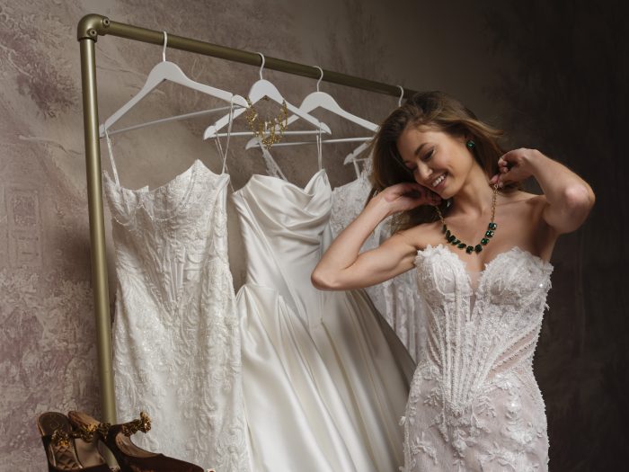 Smiling bride wearing the beaded lace Stasia gown by Maggie Sottero, fastening an emerald statement necklace and earrings, standing beside a rack of additional wedding dresses with heels in the foreground.