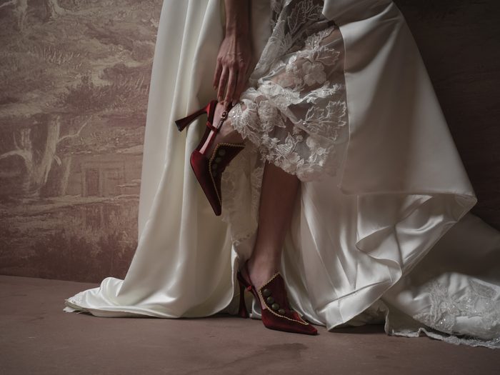 A bride's legs and hand shown fastening a red velvet button-detail heel while wearing a Maggie Sottero gown.