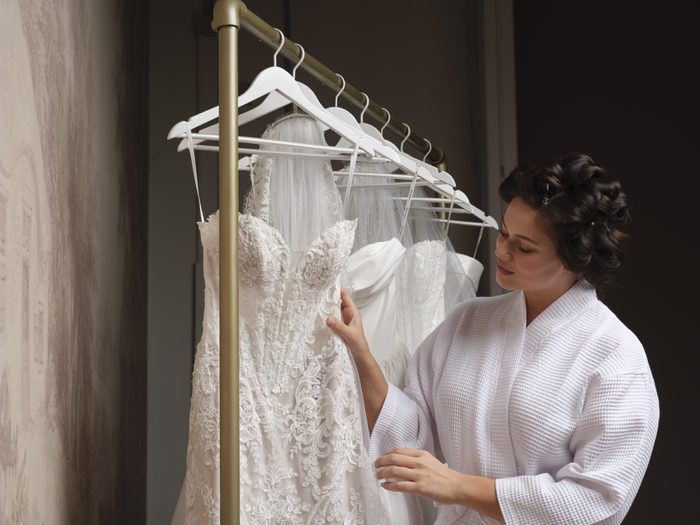 Bride in a white robe with her hair in curlers, looking at a lace wedding gown hanging on a rack alongside several other Maggie Sottero bridal gowns.