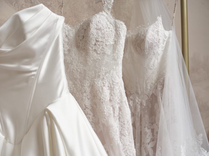 Close-up of several wedding gowns by Maggie Sottero hanging side by side on a rack, showcasing a range of fabrics and textures including smooth ivory satin, floral lace with a blush lining, and beaded lace bodices.