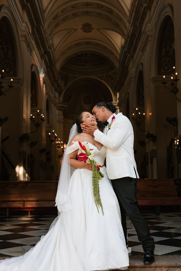 Serafina wedding gown by Maggie Sottero, an off-the-shoulder ball gown worn by a laughing bride as her groom in a white tuxedo jacket cups her face for a kiss, inside a grand cathedral with checkered marble floors and candlelit arches.