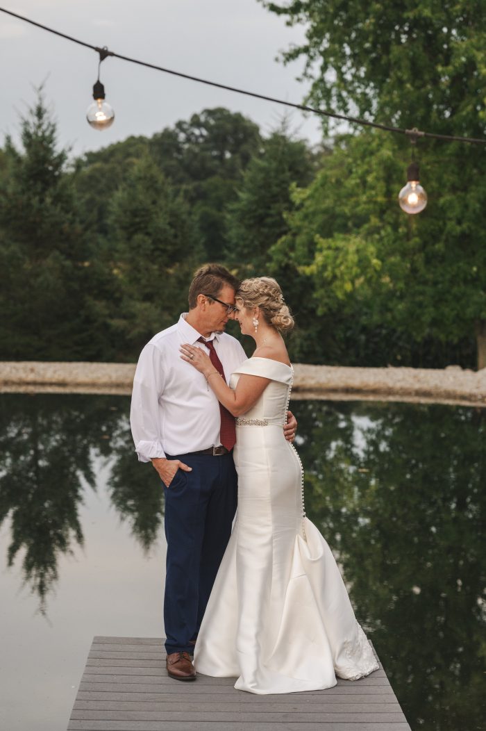 Wedding photo of a bride and groom embracing on a dock beside a pond at dusk, the bride wearing Josie by Rebecca Ingram, a fitted satin wedding dress with off-the-shoulder neckline and beaded belt.