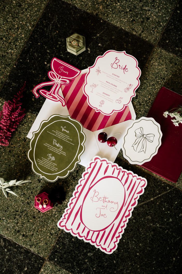 Flat lay of wedding stationery in a raspberry pink, deep red, and olive green color palette on dark speckled stone. A scalloped-edge menu card reading "Bride" lists a starter, main course, and dessert, next to a pink-and-white striped envelope with a die-cut "Groom" cocktail glass tag. An olive green card details venue, parking, and gift information. A striped invitation reads "Bethany and Joe," alongside a small card with a bow illustration, two cherries, a burgundy book, a sage velvet ring box with an engagement ring, a pink velvet earring box with pearl earrings, and pink and white florals.