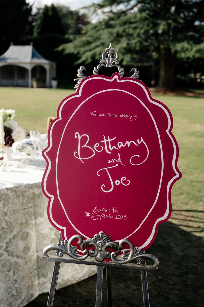 n ornate red sign with a silver decorative frame, displayed on a metal easel outdoors on a lawn. The sign reads "Welcome to the wedding of Bethany and Joe, Eaves Hall, 9th September 2025" in white cursive script. A lace-covered table with white florals and glassware is visible in the background, along with trees and a gazebo.