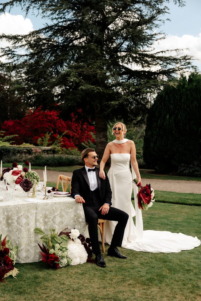 bride and groom laugh together beside an outdoor reception table styled with burgundy and white florals and lace linens. The groom, in a black tuxedo with sunglasses, sits on a wooden chair while the bride stands beside him in a strapless white gown with a draped cowl neckline, sunglasses, and a burgundy and ivory bouquet, with tall trees and red foliage in the background. Photo by Love and Luna Photography.