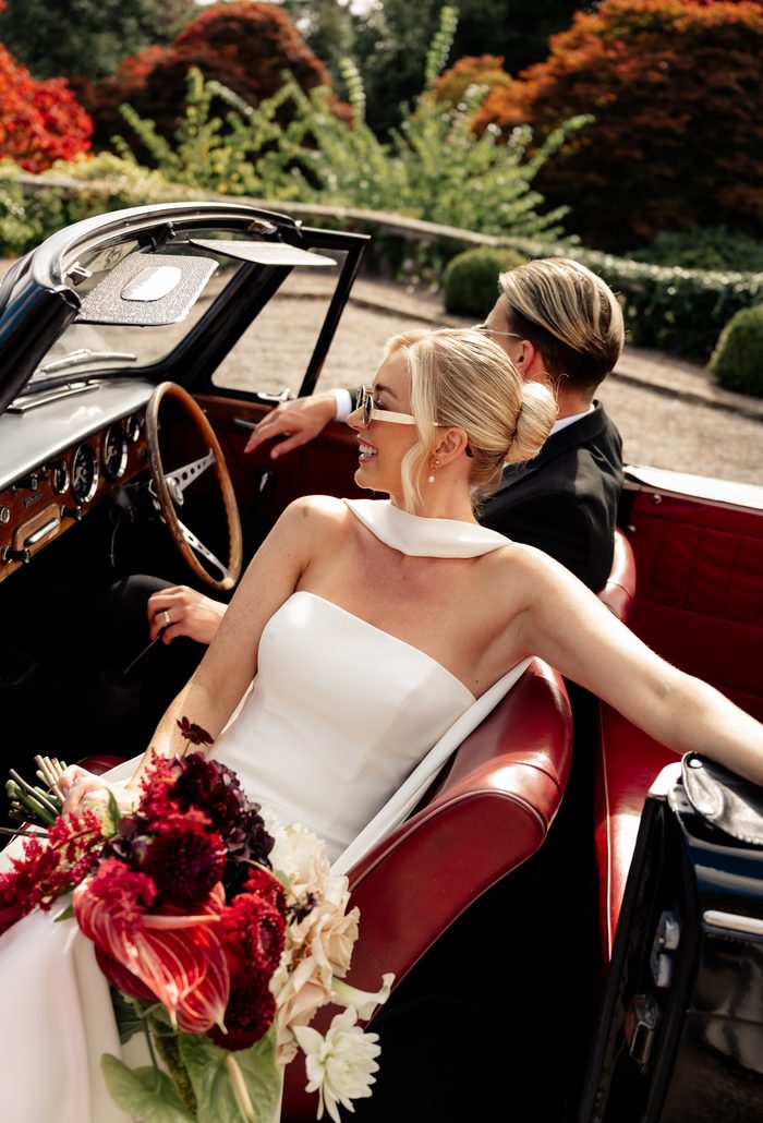 A bride and groom ride in the back of a vintage red convertible car, both wearing sunglasses and smiling. The bride, in a strapless white gown with a draped cowl neckline, holds a bouquet of burgundy and white flowers, while the groom in a black tuxedo sits beside her, with garden greenery and autumn-toned trees visible around the car. Photo by Love and Luna Photography.