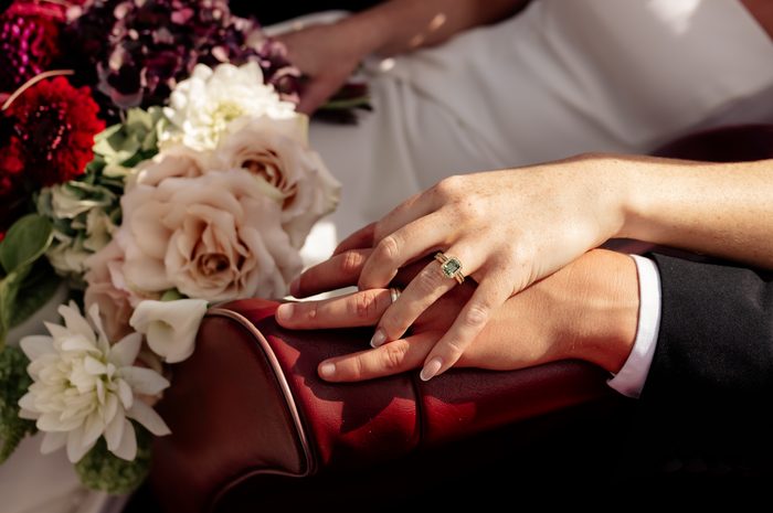 Close-up of a bride's hand resting on a groom's hand atop a burgundy leather chair arm, showing her engagement ring with a green emerald-cut stone and diamond halo alongside a wedding band. The groom wears a dark suit with a white shirt cuff. A lush bouquet of blush roses, white dahlias, deep red blooms, and dark burgundy hydrangea sits beside them, with a bride's white dress visible in the soft-focus background.