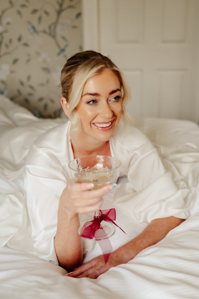 A bride smiles while lying on a white bed, wearing a white silk robe and holding a vintage coupe glass tied with a burgundy ribbon, with floral wallpaper visible softly in the background. Photo by Love and Luna Photography.