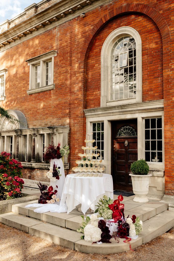 A red brick manor entrance with a stone-trimmed arched window and a wooden front door. On the steps below, a round table draped in white cloth holds a tiered champagne coupe tower with citrus garnish. Beside it, a tall floral arrangement in deep burgundy, ivory, and green sits atop a white-draped pedestal, with matching floral arrangements and greenery spilling across the stone steps.