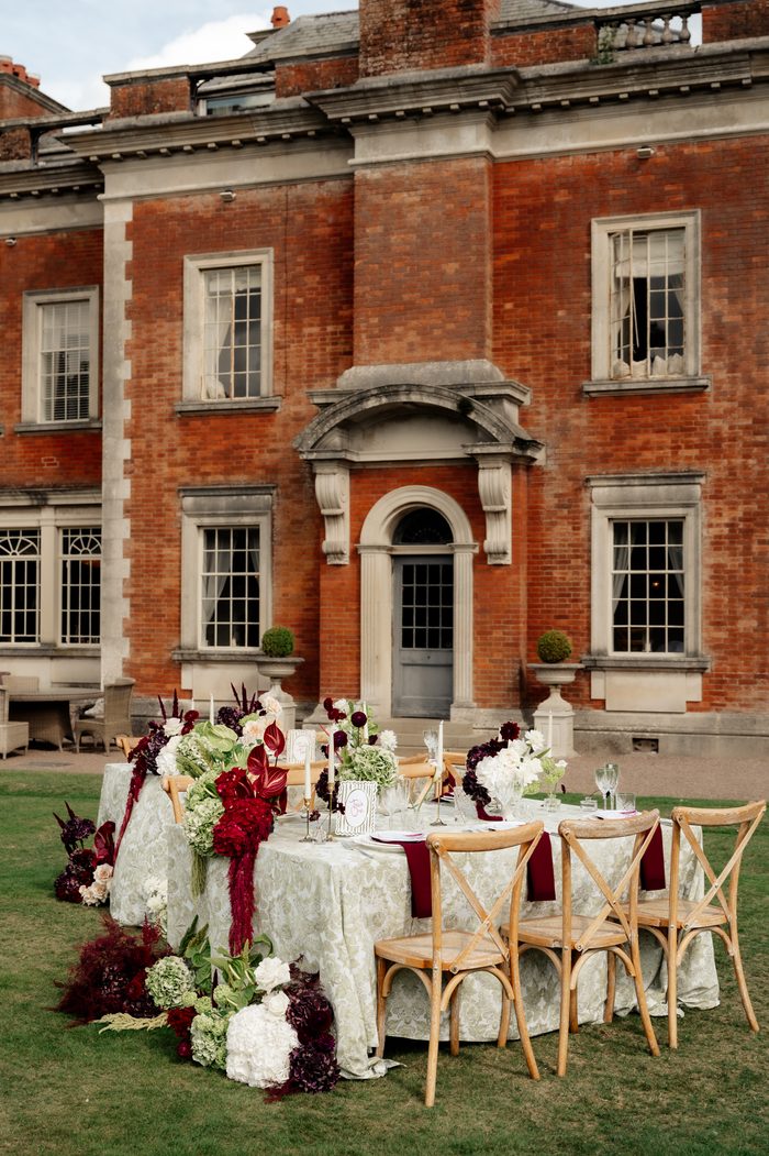 A long outdoor dining table set on a lawn in front of a grand red brick manor house with an arched stone doorway and multiple windows. The table is dressed in a light damask cloth with wooden cross-back chairs, burgundy napkins, white taper candles, and place cards. A lush floral runner of white hydrangeas, burgundy blooms, and red anthurium-like flowers cascades down the center of the table and onto the grass below.