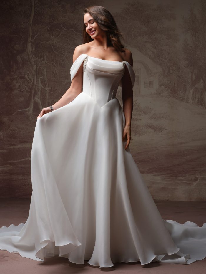 Bride wearing Abilene by Maggie Sottero, an off-the-shoulder A-line wedding dress with draped bodice, crystal detailing, and a flowing skirt with train.