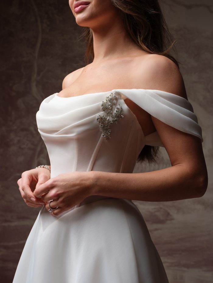 Close-up of the Abilene wedding dress by Maggie Sottero, showing the draped off-the-shoulder neckline and crystal beaded embellishment on the bodice.