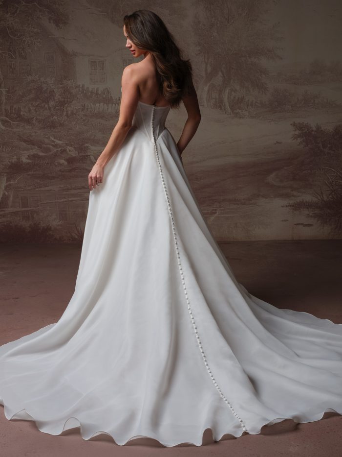 Back view of bride wearing the Abilene strapless A-line wedding dress by Maggie Sottero, featuring a fitted bodice, covered button closure, and a simple train with a wavy hem edge.