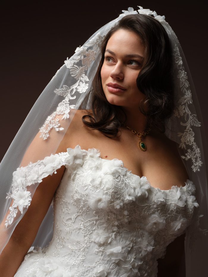 Bride wearing Benton by Maggie Sottero, a curve ballgown wedding dress with 3D floral lace bodice, off-the-shoulder cap sleeves, and matching lace-edged veil.