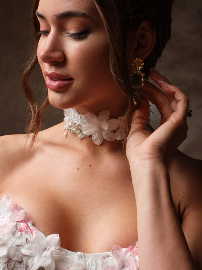 Close up of a bride wearing Benton by Maggie Sottero bridal choker, a floral lace choker with 3D floral appliqués and metallic leaf details, paired with a strapless pink floral wedding dress. 