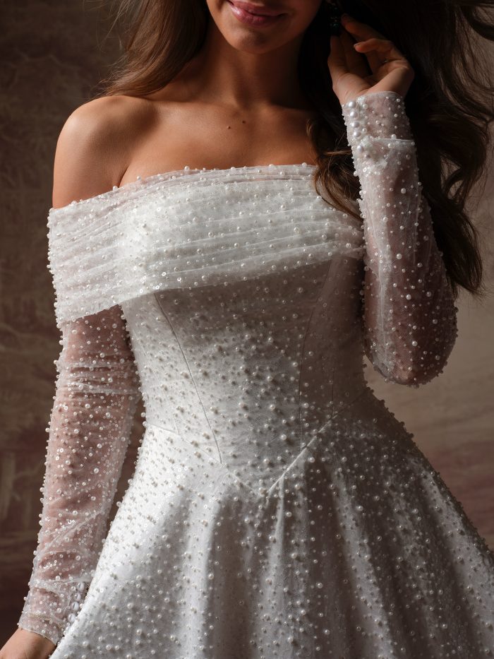 Close-up of the Elowyn A-line wedding dress by Maggie Sottero, showing allover pearl and sequin beading on the off-the-shoulder neckline, sheer long sleeves, and corset bodice.