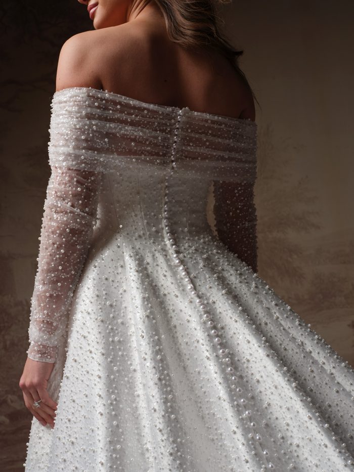 Back view of the Elowyn A-line wedding dress by Maggie Sottero, showing pearl-beaded off-the-shoulder sleeves and a corset bodice with covered button closure into a full A-line skirt.