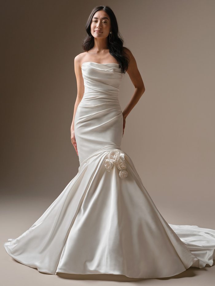 Bride wearing the Emelina fit-and-flare wedding dress by Maggie Sottero, featuring a draped satin bodice with ruching, a one-shoulder rosette accent, and a fitted mermaid skirt with a train.