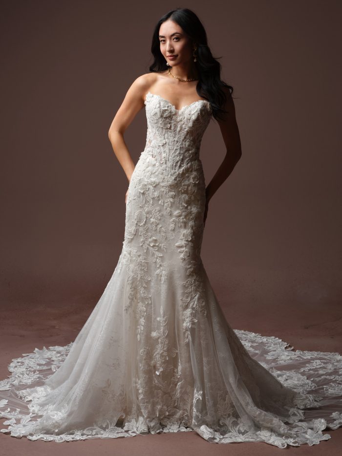 Full-length view of a bride wearing the Estelle wedding dress by Maggie Sottero, a fit-and-flare gown with a strapless floral lace bodice, sweetheart neckline, and lace-trimmed trumpet skirt with train.