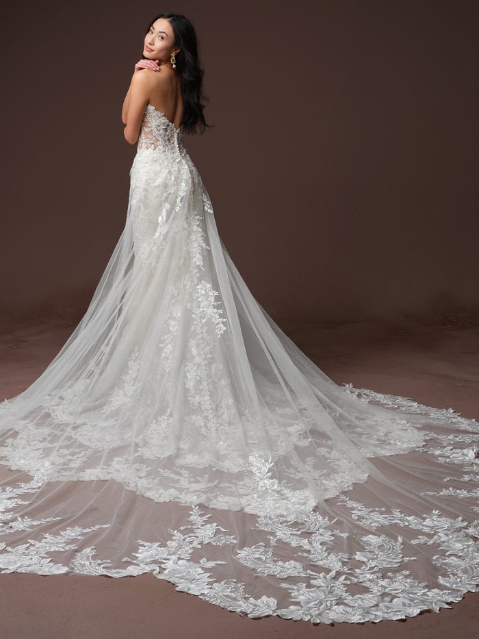 Back view of a model wearing the Estelle Fit-and-Flare Wedding Dress by Maggie Sottero in ivory, showing the fitted lace bodice with an illusion back and covered buttons, sheer lace sleeves, and a dramatic lace-edged detachable overskirt train.