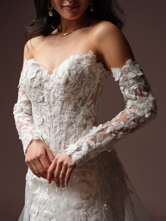 Close-up of a bride wearing the Estelle wedding dress by Maggie Sottero with detachable off-the-shoulder sleeves, showing the dimensional floral lace appliqués and beaded sweetheart bodice.