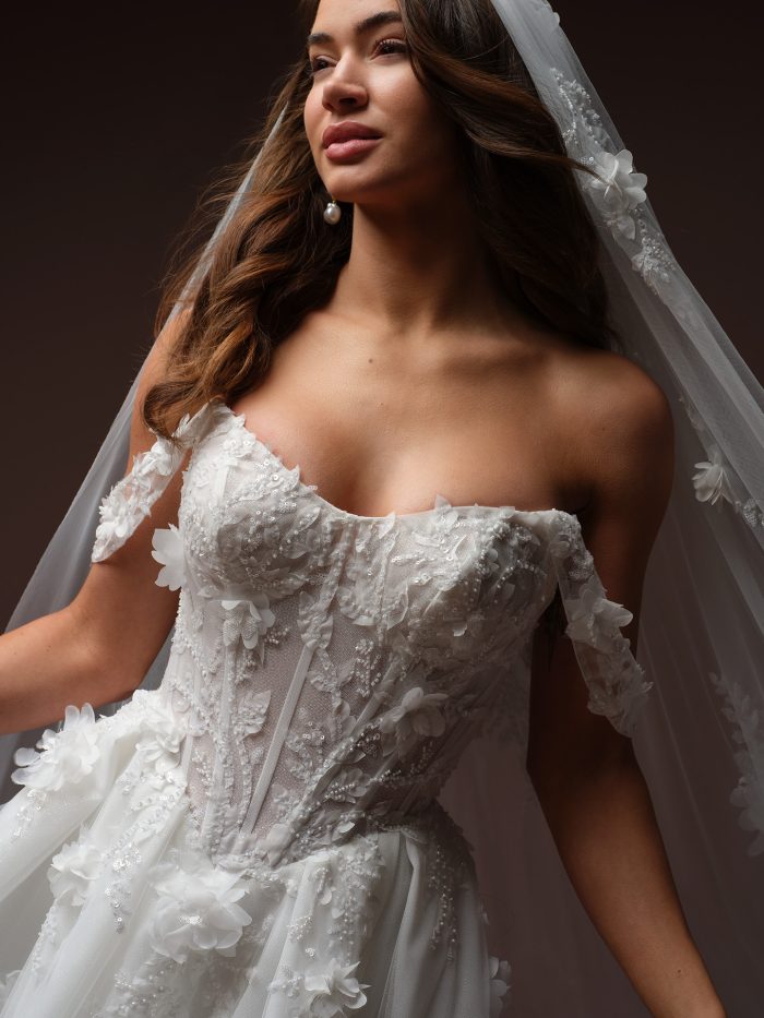 Bride wearing Fern by Maggie Sottero, an off-the-shoulder wedding dress with sequined 3D floral lace bodice, cap sleeves, and matching floral-trimmed veil.