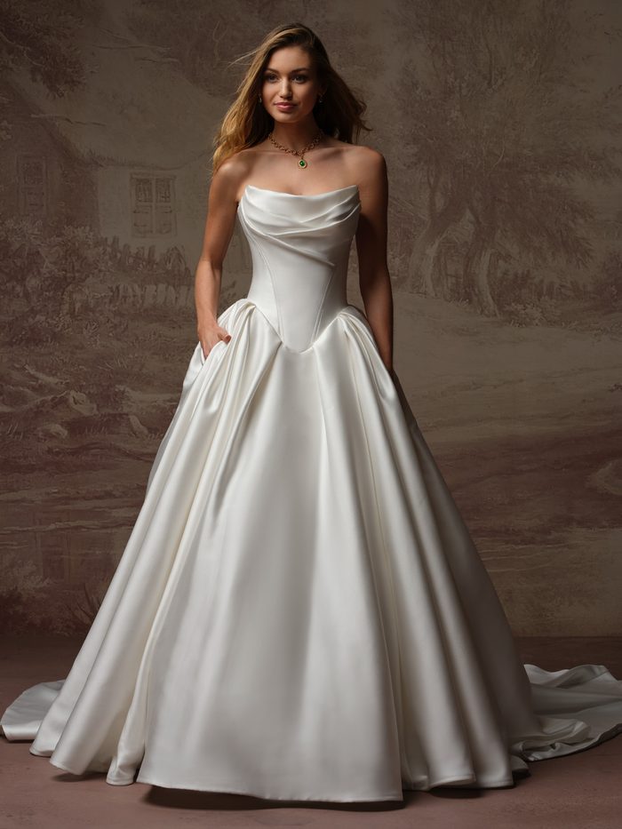 Bride wearing the Fiorita ballgown wedding dress by Maggie Sottero, featuring a draped cowl neckline, strapless satin bodice, and full pleated ballgown skirt with pockets.
