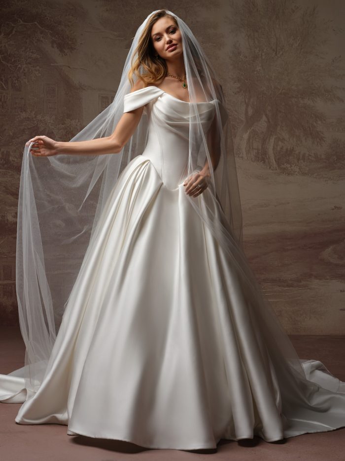 Bride wearing Fiorita by Maggie Sottero, an off-the-shoulder satin ballgown wedding dress with draped bodice and full cathedral veil.