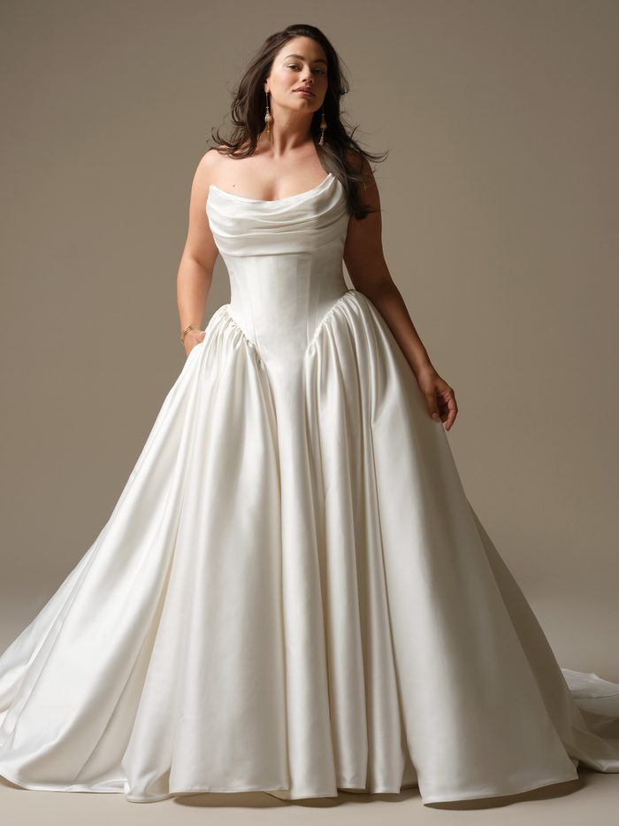 Front view of a curve plus size model wearing the Hudson Ballgown Wedding Dress by Maggie Sottero in ivory, featuring a strapless asymmetric draped neckline, a fitted structured bodice with pockets, and a full satin skirt.