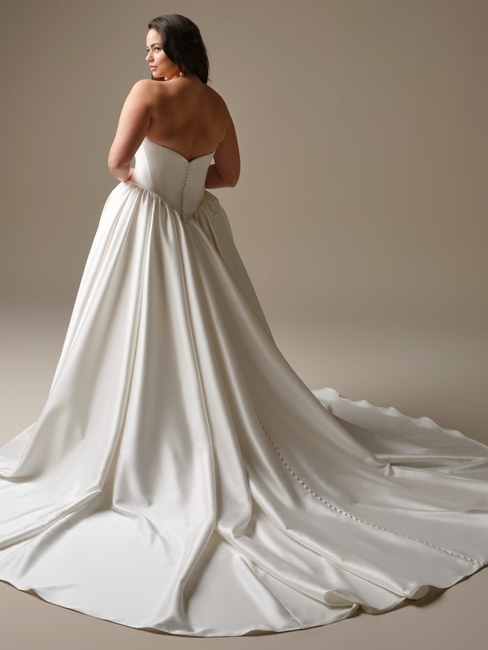 Back view of a curve plus size model wearing the Hudson Ballgown Wedding Dress by Maggie Sottero in ivory, showing the strapless bodice with a row of covered buttons down the back, and a voluminous satin ball skirt with a long train
