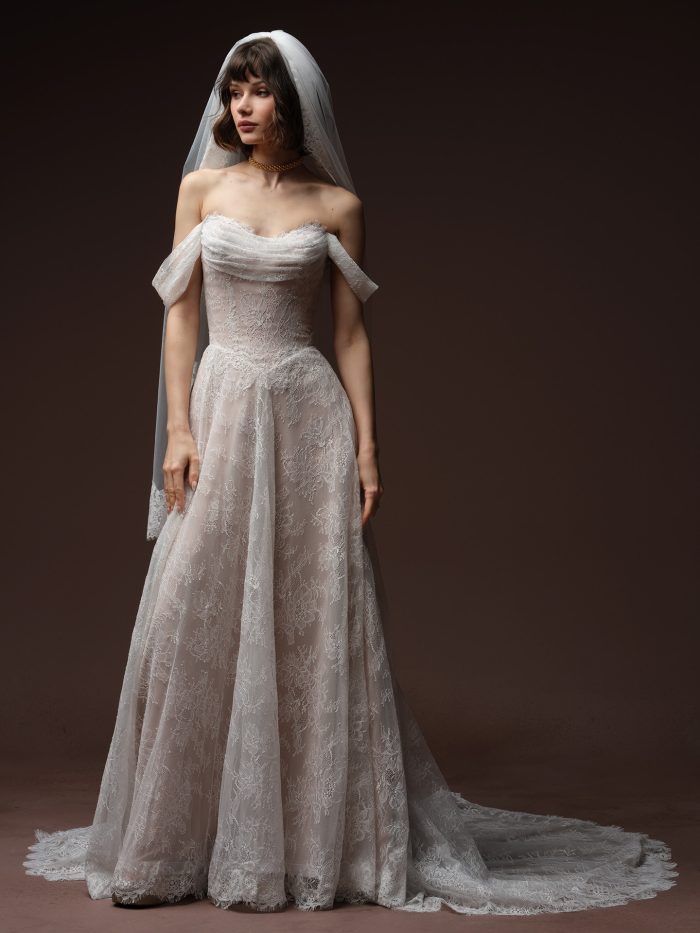 Full-length view of a bride wearing the Maelle wedding dress by Maggie Sottero, an A-line lace gown with a draped off-the-shoulder neckline and lace-trimmed veil.