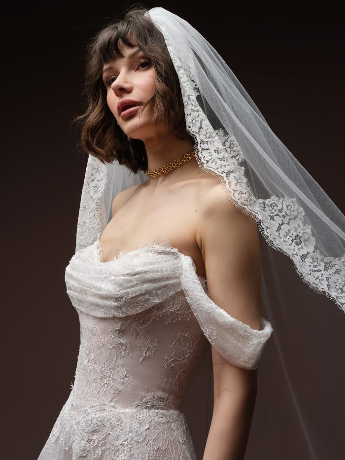 Close-up profile view of a bride wearing the Maelle wedding dress by Maggie Sottero, highlighting the draped lace off-the-shoulder bodice and scalloped lace-edged veil.