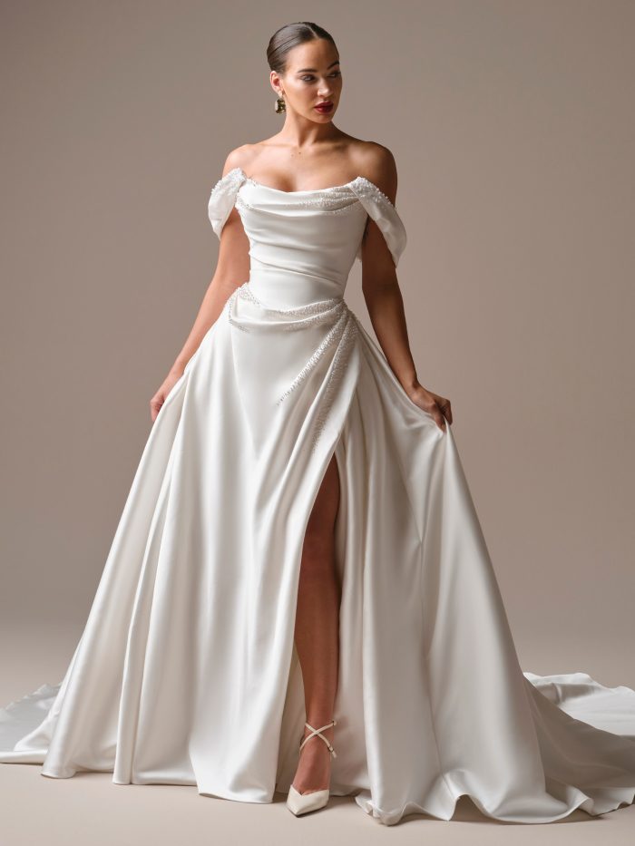 Model wearing the Kincaid wedding dress by Sottero and Midgley layered with the Melissa cap sleeve topper by Maggie Sottero, featuring a beaded off-the-shoulder cowl neckline, corset bodice, and satin skirt with front slit.