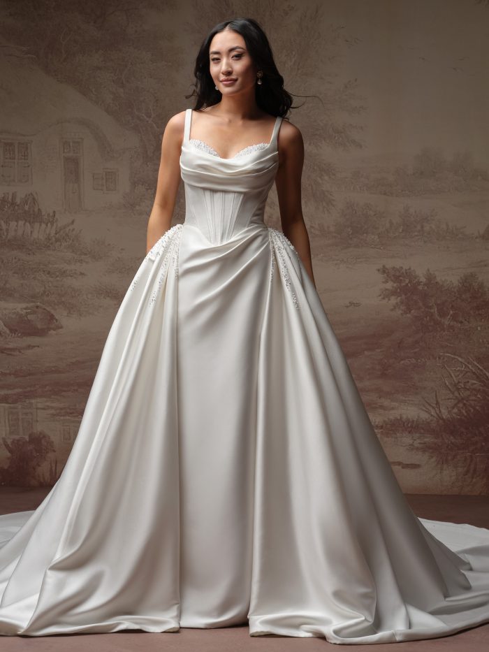 Model wearing the Melissa Royale wedding dress by Maggie Sottero, featuring a beaded cowl neckline with thin straps, corset bodice, and full satin ball gown skirt with beaded pickups.