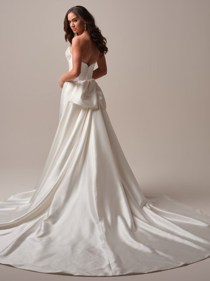 Side view of a model wearing the Melissa wedding dress by Maggie Sottero, showing the strapless corset bodice, beaded bow, and sweeping satin train.