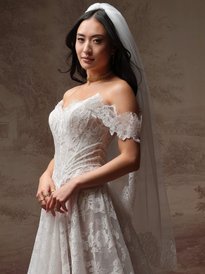 Bride wearing Mischa by Maggie Sottero, an off-the-shoulder lace A-line wedding dress with beaded waist detail and matching veil.