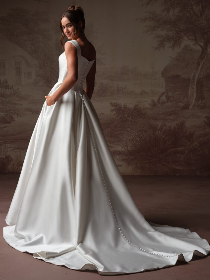 Back view of bride wearing the Pierce satin ballgown wedding dress by Maggie Sottero, featuring a square neckline with wide straps, pockets, covered button closure, and a simple train with a wavy hem edge.
