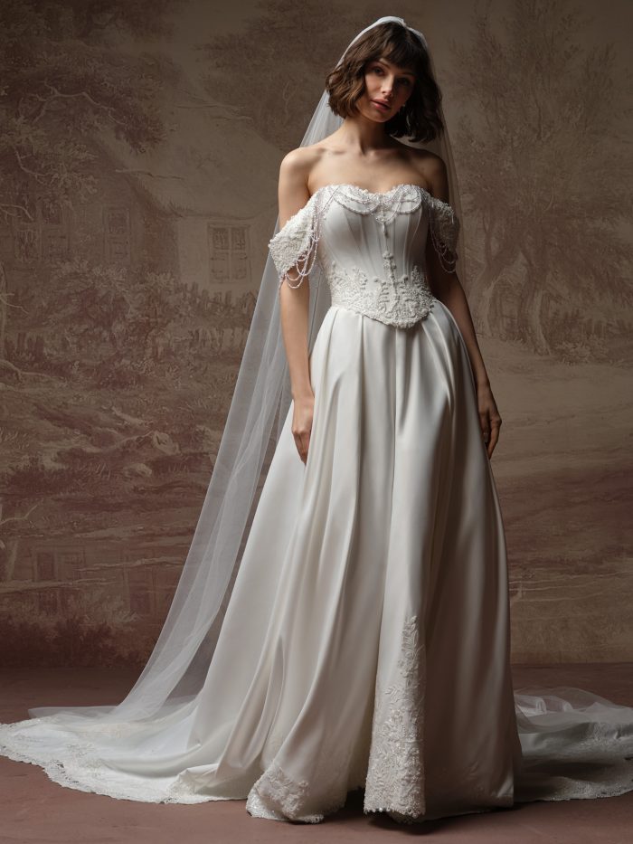 Bride wearing Seattle by Maggie Sottero, an off-the-shoulder ballgown wedding dress with corset bodice, pearl-draped cap sleeves, satin skirt with lace hem, and matching veil.