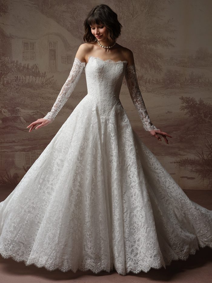 Model wearing the Serena A-line wedding dress by Maggie Sottero, featuring an off-the-shoulder lace bodice with sheer long sleeves and a full lace ballgown skirt with scalloped hem.