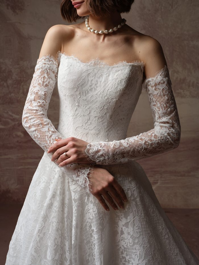 Close-up of the Serena A-line wedding dress by Maggie Sottero, showing the off-the-shoulder scalloped lace neckline and sheer lace sleeves with eyelash edging.