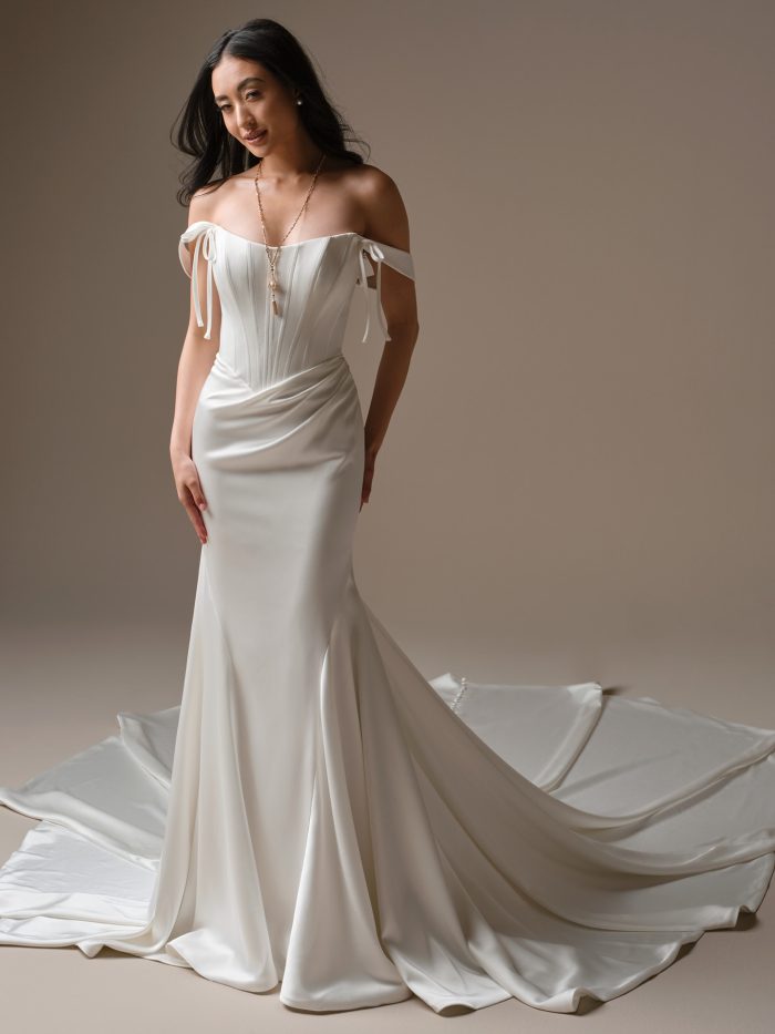 Bride wearing Sinclaire by Maggie Sottero, a fit-and-flare wedding dress with corset bodice, draped satin skirt, off-the-shoulder tie sleeves, and a dramatic train.