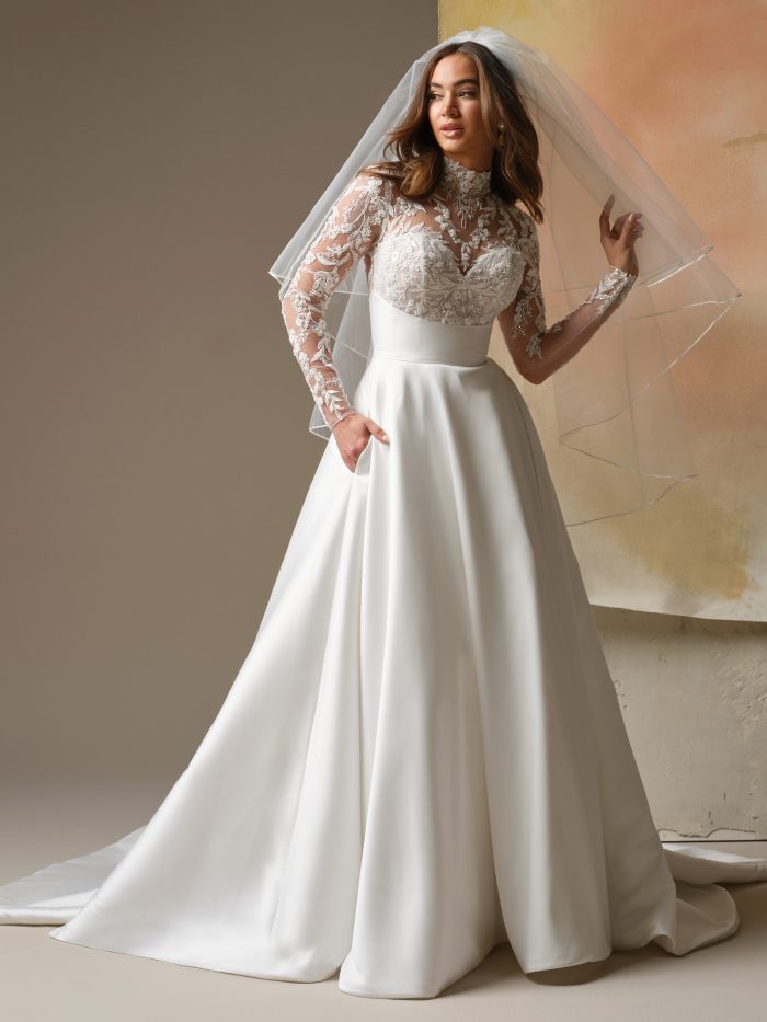 Front view of a bride wearing the Trinity A-line wedding dress by Maggie Sottero, featuring a high-neck illusion lace long-sleeve bodice, a satin waistband, and a flowing satin skirt paired with a matching veil.