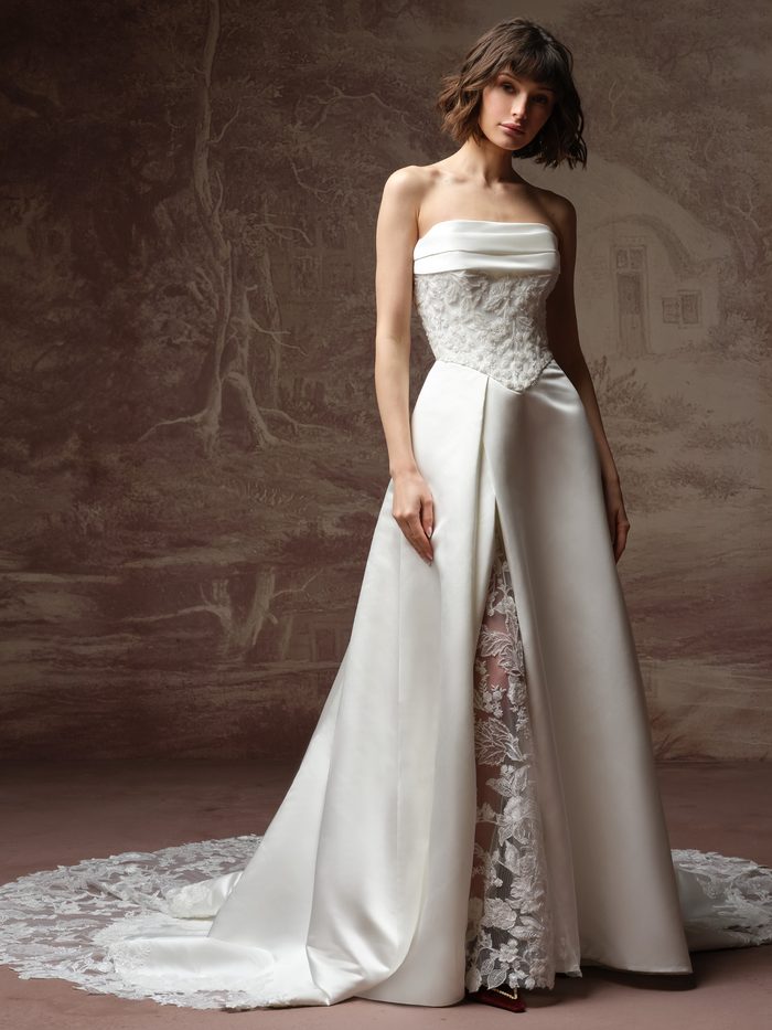 Model wearing the Wisteria A-line wedding dress by Maggie Sottero, featuring a strapless satin bodice with lace overlay, a satin skirt with front slit revealing a lace underlay, and a lace-trimmed train.