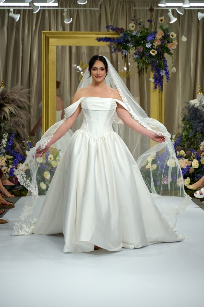 Model walking the runway in Yuma by Maggie Sottero, an off-the-shoulder ballgown wedding dress with corset bodice and lace-edged veil, shown at a Chicago bridal fashion show.