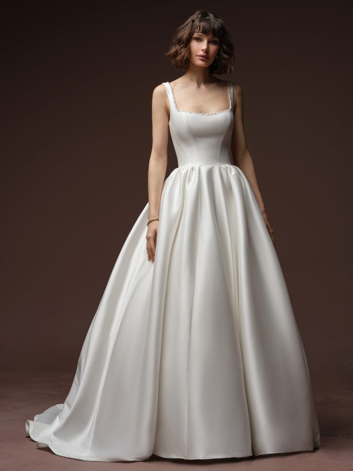 Front view of a bride wearing the Zurielle A-line wedding dress by Maggie Sottero, featuring a square neckline with beaded trim, fitted satin bodice, and a full ballgown-style skirt with a sweeping train.