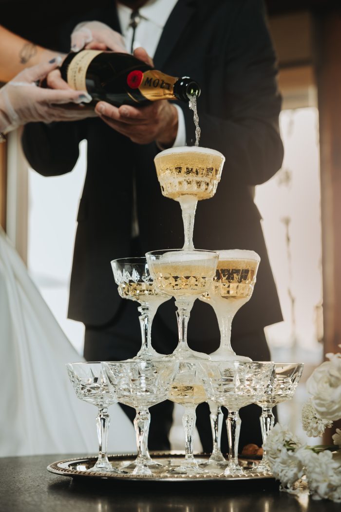 Close-up of a bride and groom pouring champagne over a tiered crystal coupe glass tower at their wedding reception, photographed by Diana Edlinger; gown by Maggie Sottero.