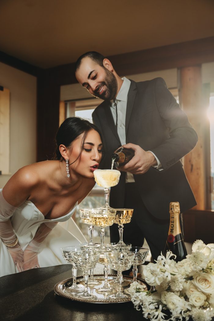 Bride sipping champagne straight from the top of a crystal coupe tower as her groom pours from a bottle of Moët, surrounded by white floral arrangements, photographed by Diana Edlinger; gown by Maggie Sottero.