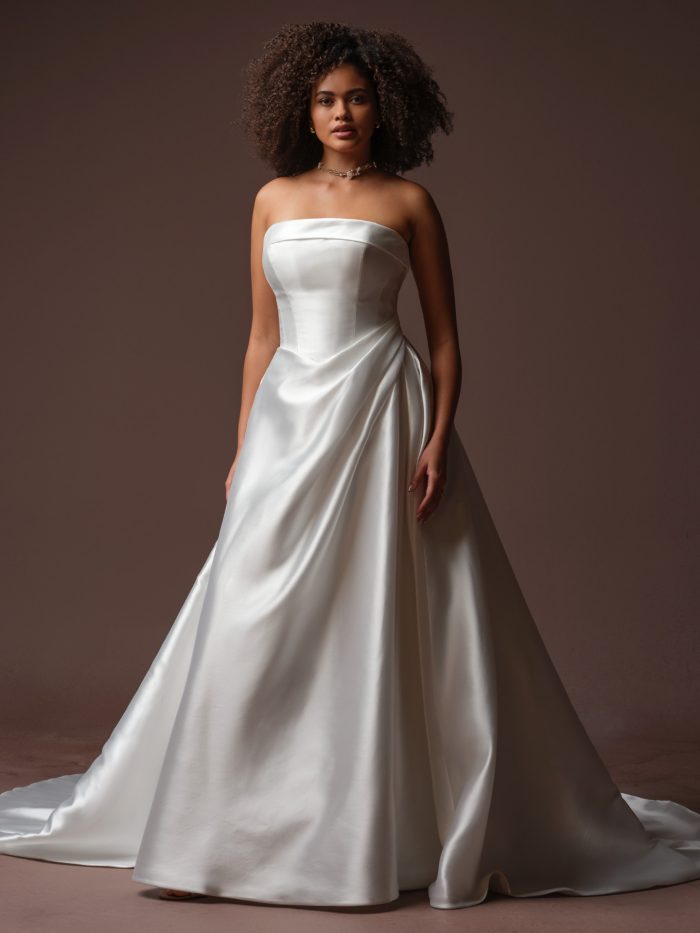 Front view of a bride wearing the Agatha A-line wedding dress by Rebecca Ingram, featuring a strapless satin bodice with draped detailing at the hip and a smooth A-line skirt with a train.