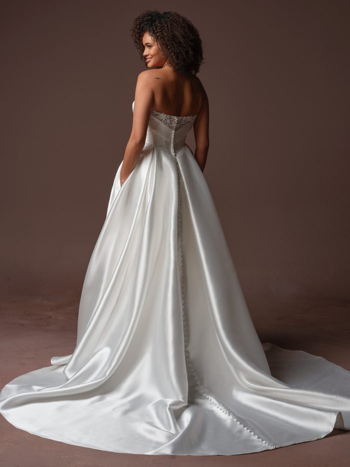 Back view of the Agatha A-line wedding dress by Rebecca Ingram, showing a strapless bodice with lace back detailing, a row of covered buttons, and a flowing satin train.