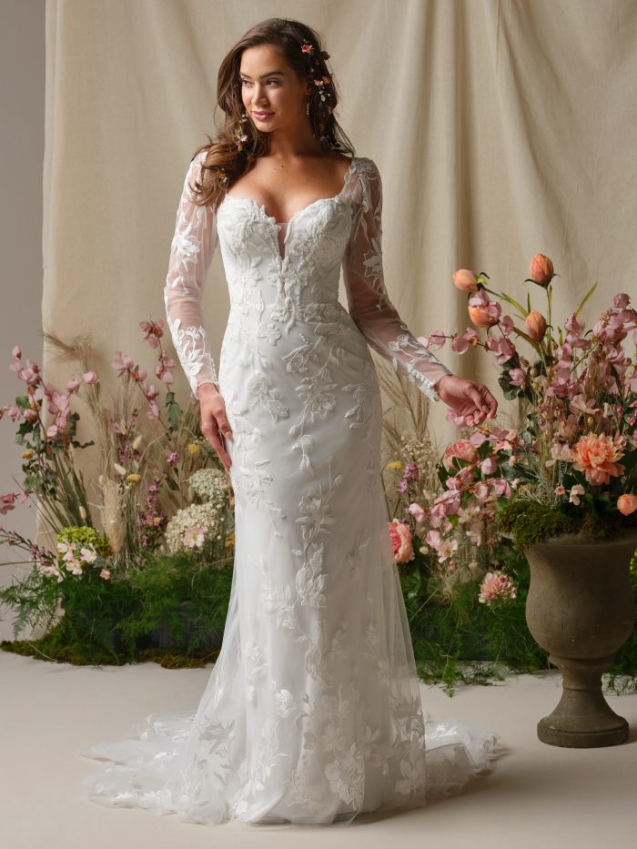 Aubra by Rebecca Ingram sheath wedding dress with floral lace appliqués, illusion long sleeves, sweetheart neckline, fitted silhouette, and soft tulle train.